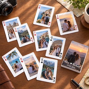 May include: A set of custom playing cards featuring various travel photographs of couples on the faces. The deck box displays a sunset mountain landscape with the printed text Alice and Michael 18.05.2026. The cards are arranged on a wooden surface.