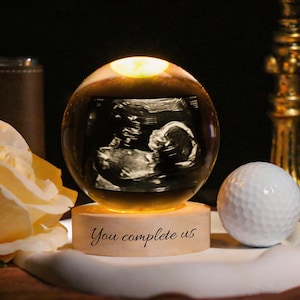 Può includere: Una sfera di cristallo luminosa con un'immagine ecografica all'interno, appoggiata su una base di legno. La base riporta la scritta "You complete us". Una pallina da golf è a destra. Lo sfondo è scuro.