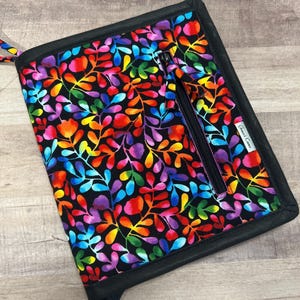 以下が含まれることがあります： 黒い背景に、虹色の鮮やかな花柄が施されたタブレットケース。ケースは黒い縁取りと黒いジッパーが付いています。お揃いの花柄のストラップが付いています。