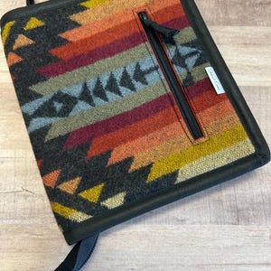 May include: A black leather-trimmed zippered pouch with a colorful geometric pattern. The pouch has a black strap and a zippered pocket on the front. The text "Great Lakes" is visible on the front of the pouch.