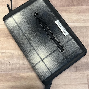 May include: A plaid zippered case in shades of gray and white, with a black border and zipper. The case has a black fabric pull and a tag that reads "Gracie's Cases". The case is rectangular and appears to be made of fabric.