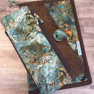 Puede incluir: Una bolsa de tela marrón y azul con cremallera que contiene una bolsa más pequeña con cremallera en su interior. La bolsa más grande tiene una solapa marrón con cierre de presión y el texto "Sewed Curves" impreso en ella.