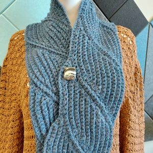 May include: A blue crocheted scarf with a silver and white decorative pin. The scarf is folded in half and draped over a brown crocheted sweater.