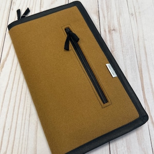 May include: A brown felt zippered pouch with a black zipper and black trim. The pouch has a small zippered pocket on the front with a black zipper pull and a black ribbon. The pouch has a black zipper pull on the bottom. The pouch has a white label with the text "Good Case" on the front.