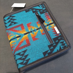 May include: A teal blue zippered notebook with a black border and a southwestern pattern. The pattern features black, red, orange, and yellow geometric shapes. The notebook has a zipper closure and a small tag that says "Made in Canada".