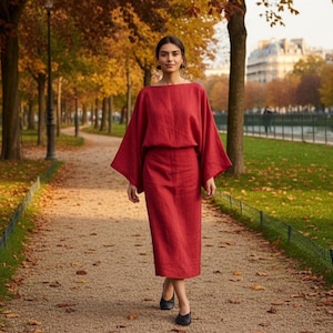 Puede incluir: Una mujer con un vestido de lino rojo, compuesto por top y falda, camina por un sendero. El vestido tiene un corte holgado y mangas anchas. El sendero está cubierto de hojas caídas, y el fondo muestra árboles y un parque.