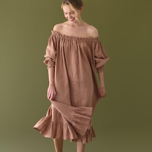 Puede incluir: Vestido marrón claro con hombros descubiertos, abotonado en la parte delantera y con dobladillo con volantes. El vestido tiene mangas largas y está hecho de una tela suave y fluida. La modelo lleva sandalias marrones. El fondo es verde oliva liso.