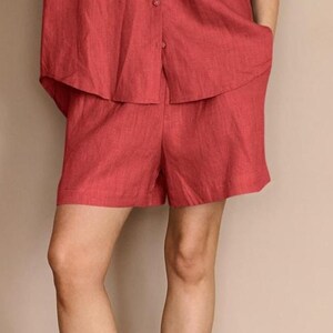 Puede incluir: Conjunto de shorts de lino rojo. El conjunto incluye una camisa con botones con dobladillo curvo y shorts a juego. Los shorts tienen bolsillos. La tela tiene un aspecto texturizado. El atuendo es informal y cómodo.