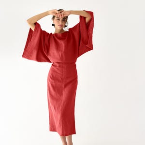 Puede incluir: Un conjunto rojo de dos piezas que consta de una parte superior con mangas anchas y una falda larga y ajustada. El conjunto está hecho de una tela ligera y se usa con bailarinas negras. Los brazos del modelo están levantados por encima de la cabeza.