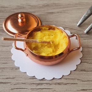 Puede incluir: Olla de cobre en miniatura con tapa, llena de comida amarilla y una pequeña cuchara de madera. La olla se encuentra sobre un mantelito de papel blanco con volantes. Dos tenedores están en el fondo. Ideal para una casa de muñecas.