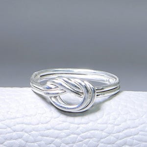 May include: A silver-colored ring with a knot design. The ring is made of a shiny metal and sits on a white surface. The knot is the focal point, with the band of the ring visible on either side of the knot.