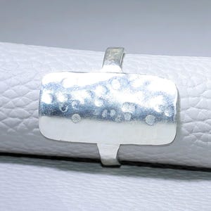 Puede incluir: Un anillo de plata con una cara rectangular y una textura martillada. El anillo tiene una banda simple y un diseño moderno. El anillo se muestra sobre una superficie blanca.