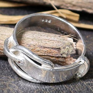 Op de afbeelding: Een zilveren ring met een uniek ontwerp, met een gebogen band en een getextureerd oppervlak. De ring is geplaatst tegen een donkere, getextureerde achtergrond met houten elementen, die de ingewikkelde details benadrukken.