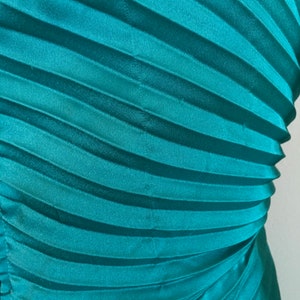 May include: Teal blue pleated fabric with a smooth, silky texture. The fabric is folded in a way that creates a series of diagonal lines.