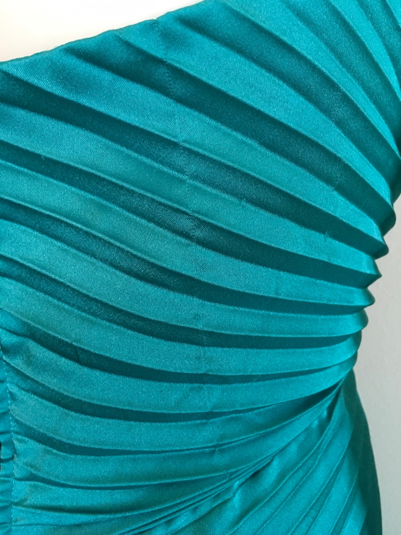 May include: Teal blue pleated fabric with a smooth, silky texture. The fabric is folded in a way that creates a series of diagonal lines.