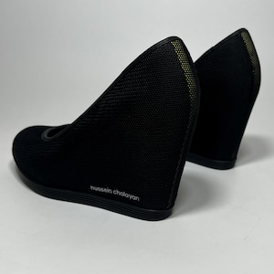 Pode incluir: Par de sapatos pretos de cunha com um design de malha texturizada. Os saltos têm bico redondo e salto cunha. O nome do designer, "hussein chalayan", está impresso na lateral do sapato.
