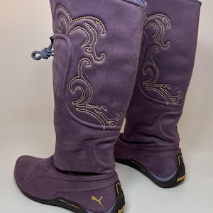 Könnte beinhalten: Ein Paar lila Wildlederstiefel mit goldfarbenen Stickereien. Die Stiefel haben einen hohen Schaft, eine runde Spitze und eine Gummisohle mit dem goldenen Puma-Logo. Die Stiefel haben einen Kordelzugverschluss oben.