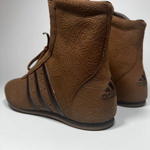 May include: Brown high-top boots with a floral embossed pattern. The boots feature the Adidas logo on the heel and three dark brown stripes on the side. The boots have brown laces and a brown sole.