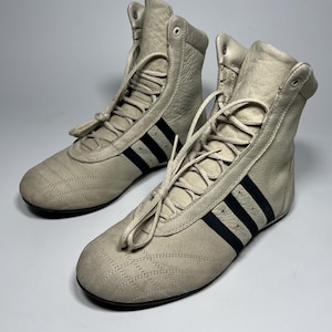 May include: A pair of high-top athletic shoes in a light beige color. The shoes feature black stripes on the sides and a lace-up closure. The toe box has a quilted design. The shoes are made of leather.