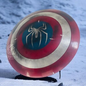 Puede incluir: Un escudo rojo, blanco y azul con un emblema de ara&ntilde;a plateada sobre un c&iacute;rculo azul. El escudo tiene un aspecto envejecido y se muestra sobre una superficie nevada. Es un accesorio o art&iacute;culo de colecci&oacute;n.