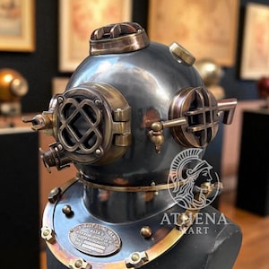 May include: A vintage diving helmet, mainly dark grey with brass detailing. The helmet has portholes, a top vent, and a neck ring with a plate that reads "MARK V". It is displayed on a black stand, highlighting its antique design and historical significance.
