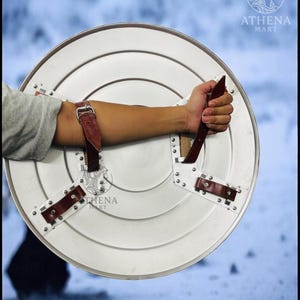 Puede incluir: Un escudo redondo de color plateado con una serie de c&iacute;rculos conc&eacute;ntricos y correas de cuero remachadas. El escudo tiene un asa y correas de cuero marr&oacute;n para asegurar al brazo. El logotipo de Athena Mart es visible.