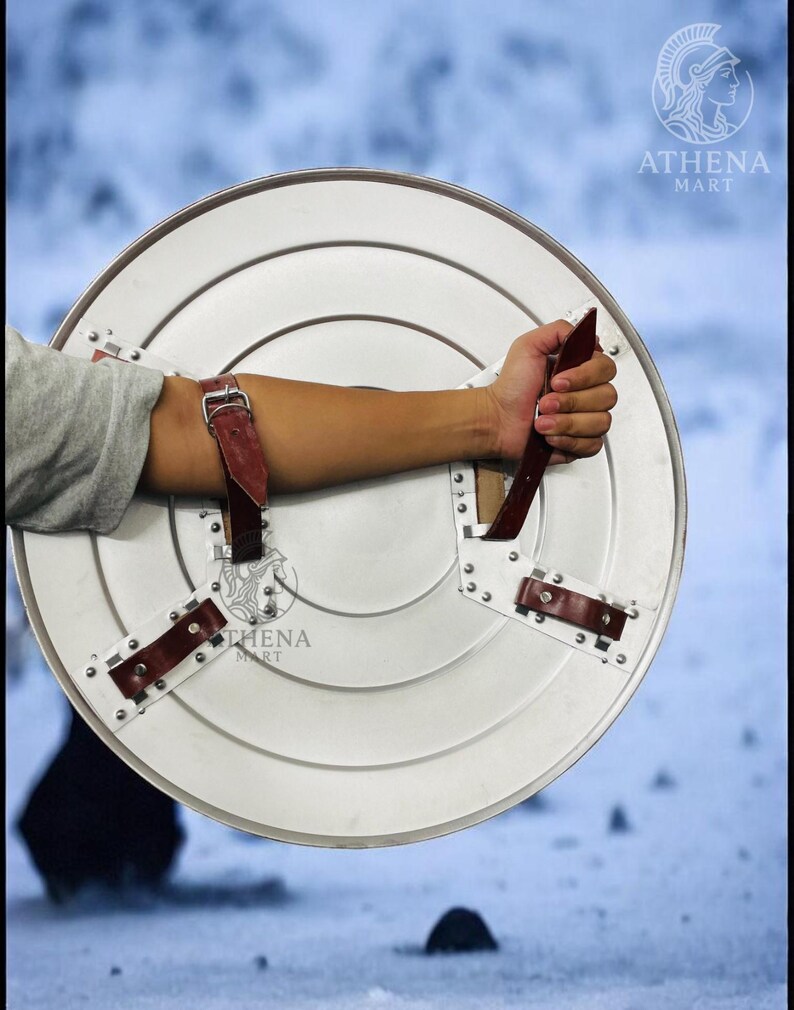Puede incluir: Un escudo redondo de color plateado con una serie de c&iacute;rculos conc&eacute;ntricos y correas de cuero remachadas. El escudo tiene un asa y correas de cuero marr&oacute;n para asegurar al brazo. El logotipo de Athena Mart es visible.