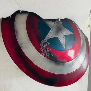 May include: A Captain America shield replica, featuring a red, white, and blue color scheme. The shield has a star in the center and is designed with a distressed, battle-worn appearance. The shield is a prop or collectible item.