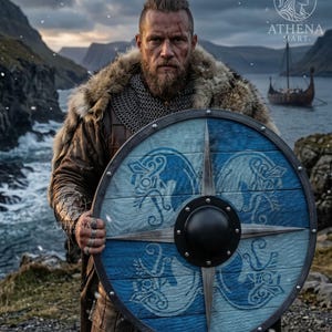 Peut inclure: Un bouclier viking rond avec un motif bleu et blanc, avec un bossage noir central et une rose des vents stylisée. Le bouclier a un aspect en bois avec un bord en métal. L'image montre également un guerrier viking en tenue traditionnelle.