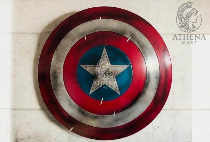 May include: A round shield with a red, white, and blue color scheme. The shield features a central blue circle with a white star, surrounded by red and white concentric rings. The shield has a distressed, weathered appearance, and the Athena Mart logo is visible.