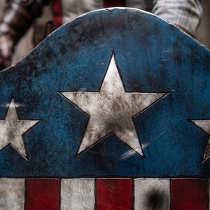 May include: A distressed blue, white, and red shield with three large white stars and red and white stripes. The shield has a weathered appearance, suggesting it is a replica or prop. The shield is a symbol of heroism and patriotism.
