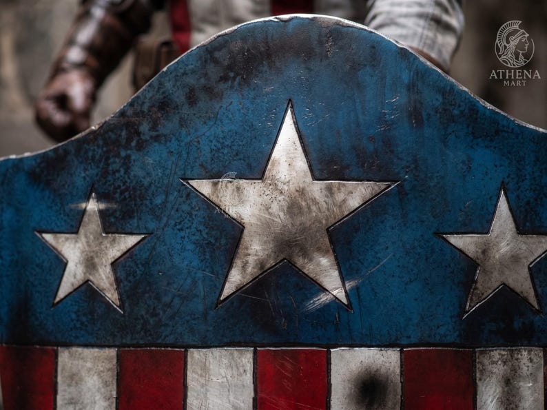May include: A distressed blue, white, and red shield with three large white stars and red and white stripes. The shield has a weathered appearance, suggesting it is a replica or prop. The shield is a symbol of heroism and patriotism.