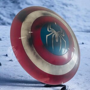 Puede incluir: Un escudo rojo, blanco y azul con un emblema de ara&ntilde;a plateada en el centro. El escudo tiene un aspecto desgastado y est&aacute; sobre un fondo nevado. Es un accesorio o art&iacute;culo de colecci&oacute;n.