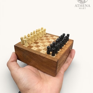 Op de afbeelding: Een kleine houten reisschaakset met een dambordpatroon. De schaakstukken zijn zwart en crèmekleurig. De set wordt in een hand gehouden. De houten doos is donkerbruin. Het Athena Mart-logo staat in de rechterbovenhoek.
