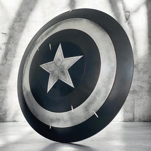 K&ouml;nnte beinhalten: Ein Captain America-Schild mit einem zentralen wei&szlig;en Stern auf einem blauen Kreis, umgeben von konzentrischen silbernen und schwarzen Ringen. Der Schild ist rund und hat ein verwittertes, metallisches Aussehen mit sichtbaren Kratzern.