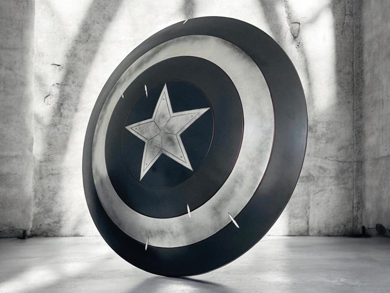 K&ouml;nnte beinhalten: Ein Captain America-Schild mit einem zentralen wei&szlig;en Stern auf einem blauen Kreis, umgeben von konzentrischen silbernen und schwarzen Ringen. Der Schild ist rund und hat ein verwittertes, metallisches Aussehen mit sichtbaren Kratzern.