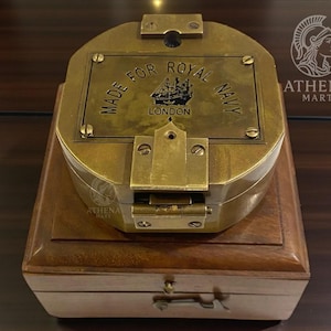 Peut inclure: Une boussole en laiton vintage, marquée "MADE FOR ROYAL NAVY LONDON", est posée sur une boîte en bois. La boussole a un couvercle à charnière et est fixée avec des vis. La boîte a un petit tiroir. La boussole est de couleur or antique.