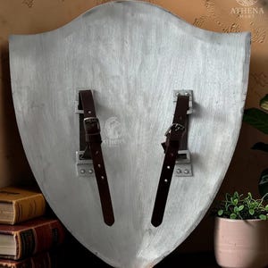 May include: A silver-colored shield with two brown leather straps and buckles. The shield has a slightly textured surface and a curved shape. The back of the shield is visible, showing the mounting hardware.