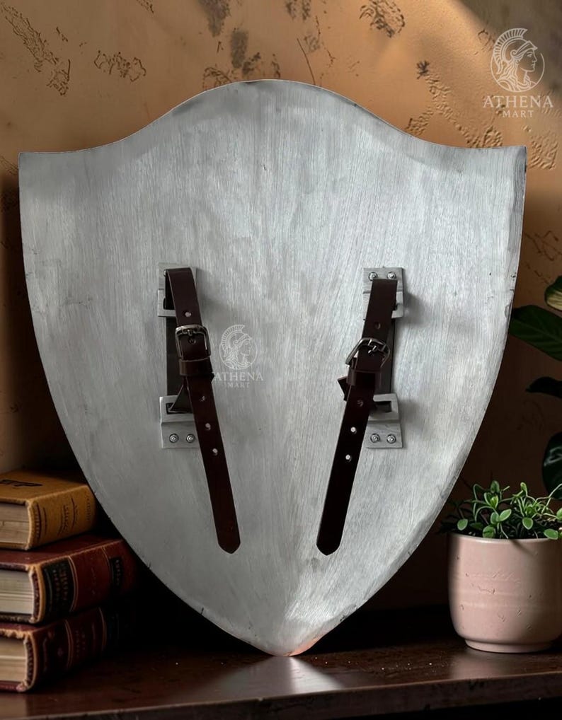 May include: A silver-colored shield with two brown leather straps and buckles. The shield has a slightly textured surface and a curved shape. The back of the shield is visible, showing the mounting hardware.