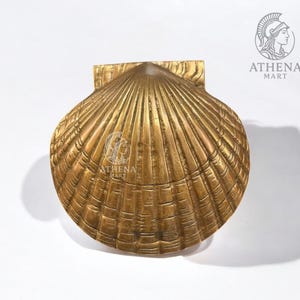 Peut inclure: Un objet décoratif en forme de coquillage de couleur dorée. L'objet présente une surface texturée avec des nervures détaillées et une plaque de montage rectangulaire. Le logo Athena Mart est visible sur l'objet.