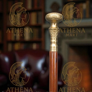 Pode incluir: Uma bengala de madeira com cabo e detalhes em latão. A pega tem um design ornamentado e um topo arredondado. O eixo da bengala é de cor castanho escuro. O logótipo Athena Mart é visível no fundo.