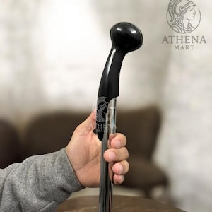 Könnte beinhalten: Ein schwarzer, gebogener Gehstock mit rundem Griff. Der Griff und der gebogene Teil sind schwarz, während der Schaft eine glänzende silberne Farbe hat. Der Stock wird von einer Person gehalten, die einen grauen Pullover trägt.