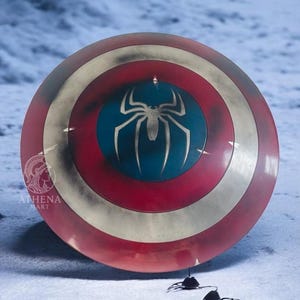 Puede incluir: Un escudo redondo con una combinaci&oacute;n de colores rojo, blanco y dorado. El centro presenta un c&iacute;rculo azul con un emblema de ara&ntilde;a blanco. El escudo es un accesorio o art&iacute;culo de colecci&oacute;n, probablemente inspirado en un tema de superh&eacute;roes.