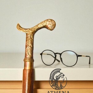May include: A decorative walking cane with a detailed gold-colored handle and a wooden shaft. Round black eyeglasses rest on a white surface. The Athena Mart logo is visible.