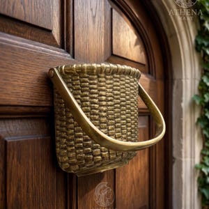 Puede incluir: Un llamador de puerta en forma de cesta de color bronce, con un asa curvada, montado en una puerta de madera oscura. El llamador tiene una textura tejida y está diseñado para parecerse a una pequeña cesta. La puerta es de madera oscura.