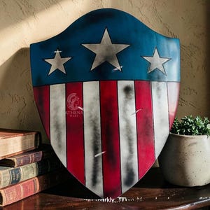 May include: A shield with a blue top section featuring three white stars and a red and white striped bottom. The shield has a distressed, vintage appearance. The Athena Mart logo is visible on the shield. The shield is displayed on a wooden surface.