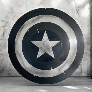 K&ouml;nnte beinhalten: Ein gro&szlig;er, runder Schild in Schwarz, Wei&szlig; und Silber. Der Schild hat einen zentralen wei&szlig;en Stern, der von konzentrischen Kreisen umgeben ist. Dieser Schild ist eine Nachbildung von Captain Americas Schild.