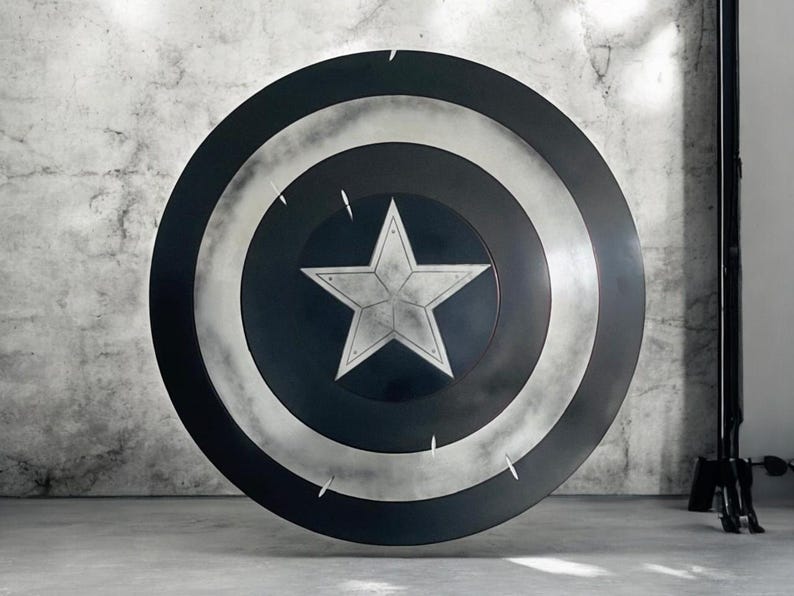 K&ouml;nnte beinhalten: Ein gro&szlig;er, runder Schild in Schwarz, Wei&szlig; und Silber. Der Schild hat einen zentralen wei&szlig;en Stern, der von konzentrischen Kreisen umgeben ist. Dieser Schild ist eine Nachbildung von Captain Americas Schild.
