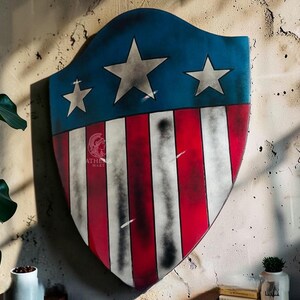 May include: A shield with a blue top section featuring three white stars and a red and white striped lower section. The shield is mounted on a textured wall. The shield has a distressed, vintage appearance.