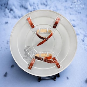 Puede incluir: Un escudo redondo de color plateado con correas de cuero marr&oacute;n y remaches. El escudo tiene un borde ligeramente elevado y un dise&ntilde;o central. El logotipo "ATHENA MART" es visible en el escudo.